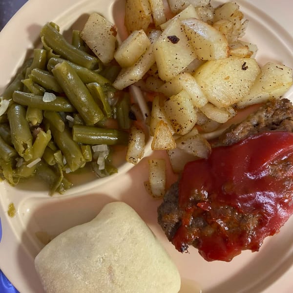A plate of American cuisine featuring meatloaf with ketchup, green beans, fried potatoes, and a roll.