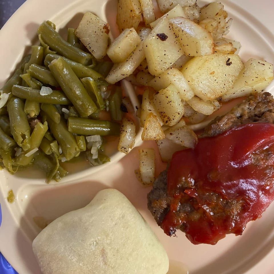 A plate of American cuisine featuring meatloaf with ketchup, green beans, fried potatoes, and a roll.