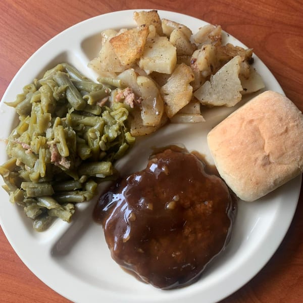 American Salisbury steak with gravy, green beans, roasted potatoes, and a dinner roll on a white plate.